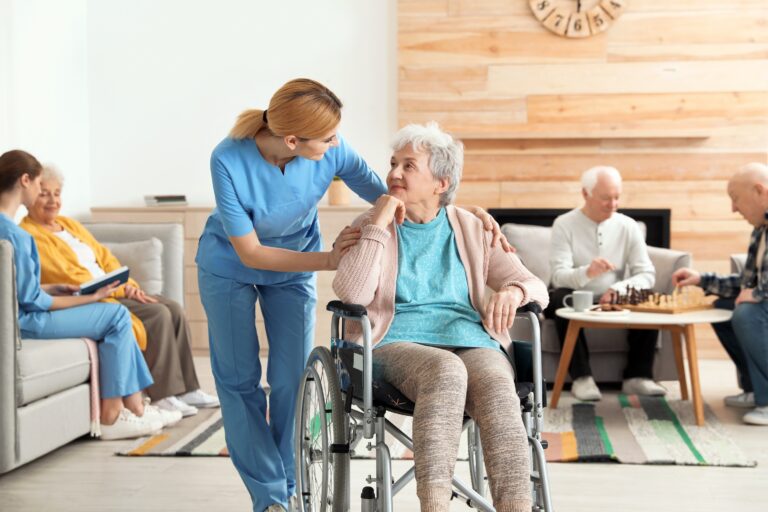 Nurse in blue scrubs assists an elderly woman in a wheelchair, smiling and talking in a bright common room, others seated nearby.