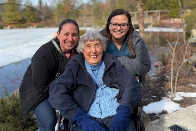 Three women pose outdoors in winter: an elderly woman in a wheelchair sits center, flanked by two younger women, all smiling with a snowy lake and trees in the background.