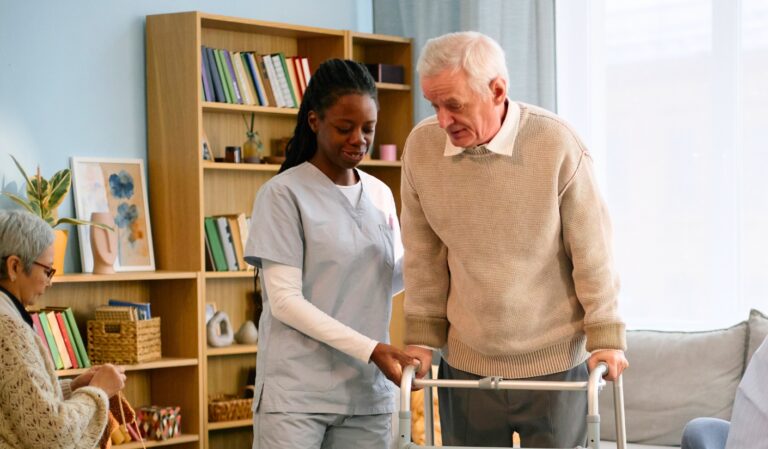 Caregiver helps an elderly man using a walker in a bright living room while another senior knits in the background.
