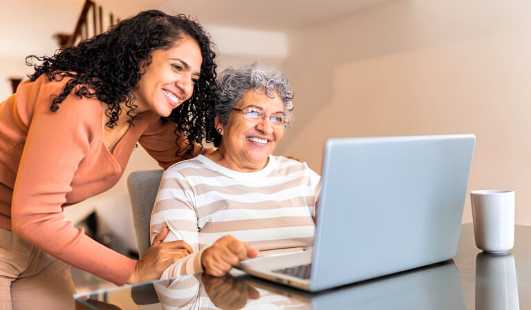 Two women, one younger and one older, smile at a laptop on a glass table as they work together.