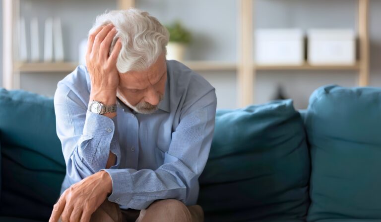 Older man with gray hair sitting on a blue sofa, head resting in one hand, looking stressed or troubled.