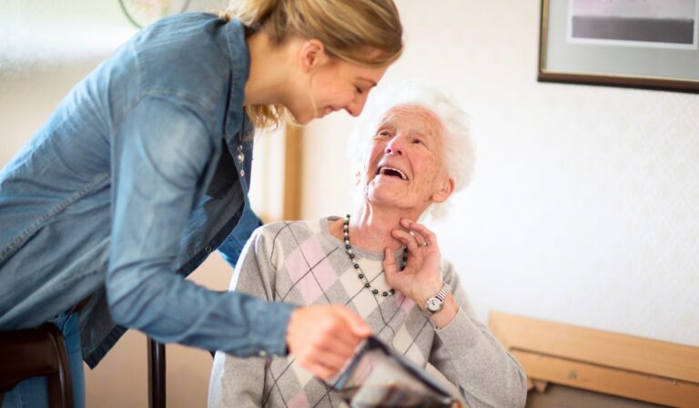 Caregiver leans toward an elderly woman who smiles warmly as they share a moment together indoors.