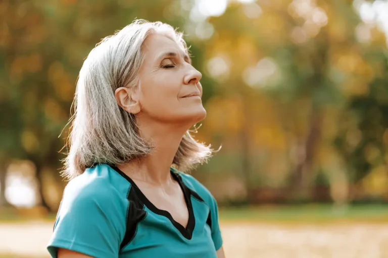 Older woman with gray hair wearing a teal athletic shirt, standing outdoors with eyes closed in a peaceful pose.